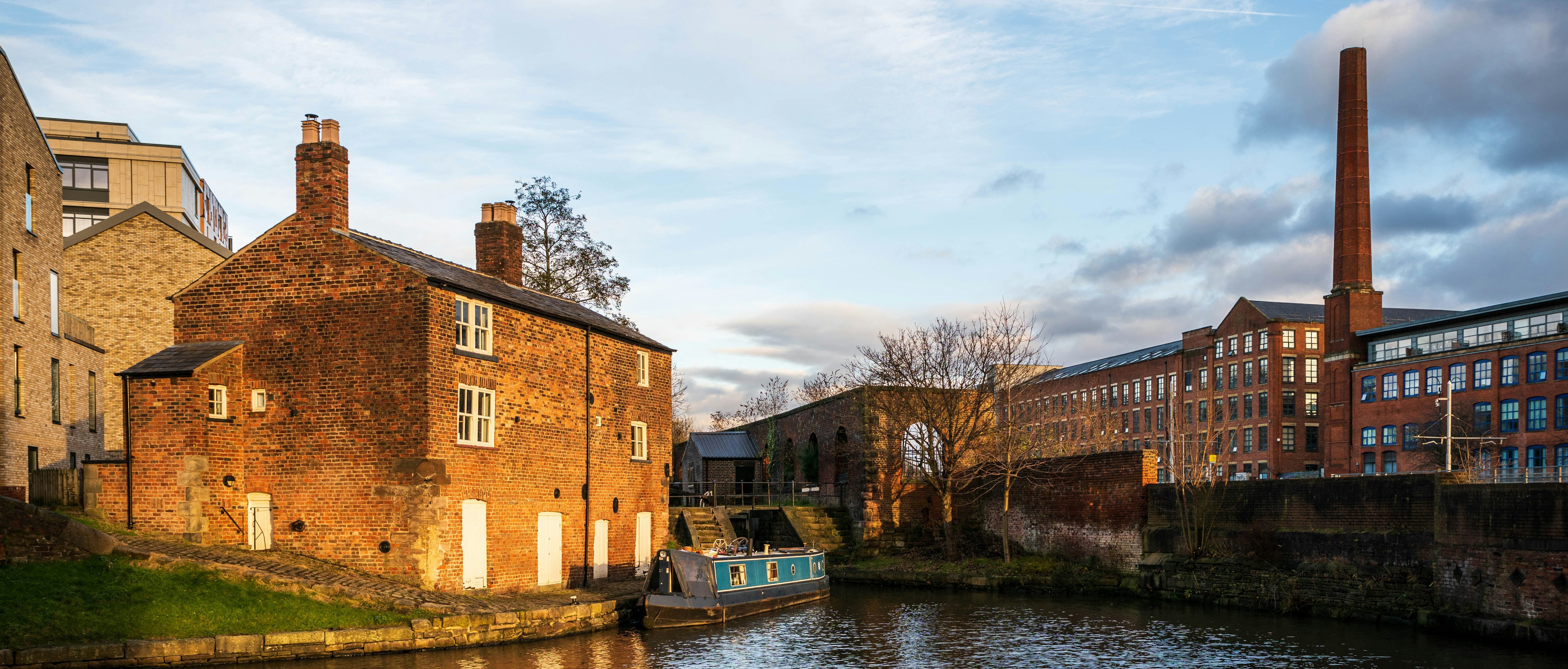 An image of a narrowboat moored in an urban area. A mill with a chimney stack is visible in the background. Image by Jason Jeandron on Unsplash
